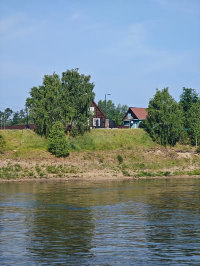 The image shows a waterfront scene in Galanino, Russia, with two houses situated on a grassy hill overlooking a wide river. The house on the left is a dark brown wooden structure with white-trimmed windows and a dark door. A large birch tree with lush green foliage stands prominently in front of it. The house on the right is smaller, painted a light teal color with a reddish-brown roof. It has white-trimmed windows and a balcony. The hill is covered in green grass with patches of sandy soil visible. The river in the foreground is calm, reflecting the sky and surrounding greenery. The sky is clear and blue with a few wispy clouds. There is a brown fence running along the top of the hill behind the houses, and a street lamp is visible to the left of the teal house. A road sign with a yellow triangle is also visible in the distance to the left.