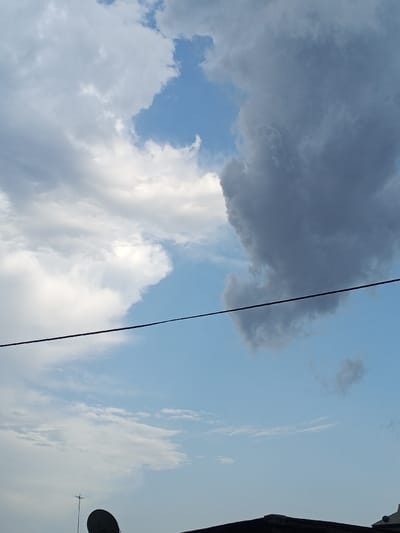 The image depicts a sky view over Konongo, Ghana. The upper half of the frame shows a dynamic cloudscape, with patches of clear blue sky interspersed among white cumulus clouds and a large, dense, dark grey cloud formation occupying the upper right quadrant and extending towards the center. A single, thin, dark horizontal wire or cable traverses the mid-ground. The lower portion of the image reveals elements of a rooftop: a dark, partial satellite dish is visible in the bottom left, alongside a thin, vertical antenna. To the right, a dark, angular roofline is present, with a lighter-toned structure partially visible on the far bottom right. No individuals or active interactions are observable.