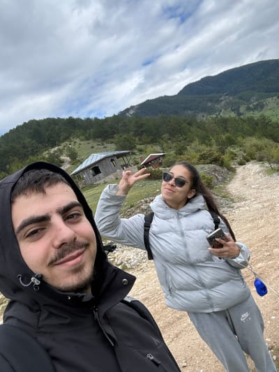 Two individuals pose for a selfie. A male figure is centered in the foreground, wearing a black hooded jacket. To the left, a female figure stands behind the male, wearing a light grey puffer jacket and grey sweatpants. She is holding a cell phone in her left hand and gesturing with a peace sign in her right hand. Both individuals are looking at the camera.

The background reveals a partly cloudy sky, a wooded mountain range, and a small wooden building situated on a grassy hillside. The location is Giovren, Bulgaria.