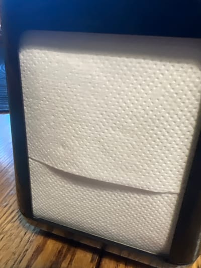 The image shows a close-up view of a napkin holder filled with white paper napkins. The napkin holder appears to be made of dark material, possibly wood or plastic, and is placed on a wooden table with a visible grain. The napkins have a textured surface with small embossed dots. A slight shadow is cast on the napkins from the top of the holder. The background is dark and out of focus, with a hint of blue. The scene is indoors, on a table likely in a restaurant or dining setting. The image does not contain any people or discernible actions. There is no information within the image to suggest a specific location in Berehove, Ukraine.