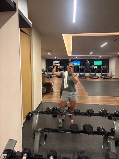 In a gym mirror selfie, a person flexes a bicep. A gray t-shirt, light-colored shorts, and a white towel draped over a shoulder are worn. Dumbbells stand to the right. Treadmills with televisions are visible in the background. The scene is brightly lit, suggesting a modern fitness center.