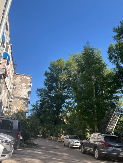 The image depicts an outdoor scene on a clear, sunny day in the city of Kiselyovsk, Russia. In the foreground, a silver car is parked on the left side of a paved road. To its right, a dark gray SUV is also parked. Beyond these vehicles, a narrow street continues, lined with trees on the right and apartment buildings on the left. The apartment buildings appear to be older Soviet-era structures, with one on the left showing signs of wear and balconies with blue trim. Large, leafy trees dominate the right side of the frame, casting shadows on the road. In the background, more trees and buildings are visible. The sky is a vibrant, cloudless blue.