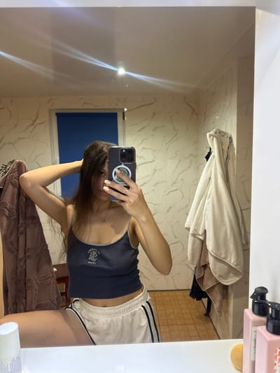 A young woman is taking a mirror selfie in a bathroom. She is wearing a dark blue tank top with a graphic print and white shorts with black stripes. Her hair is long and dark, and she is holding it up with one hand. She is wearing a cross necklace and a ring on her finger. In the mirror, we can see a blue-tiled wall with marble-patterned accents, a blue roller blind on a window, and several towels hanging on a rack. There are also pink bottles with black pumps on the counter in the foreground. The flooring appears to be tiled in a light brown color. The overall impression is a casual, domestic setting. There is no visual information in the image that would allow for the identification of the city or country.