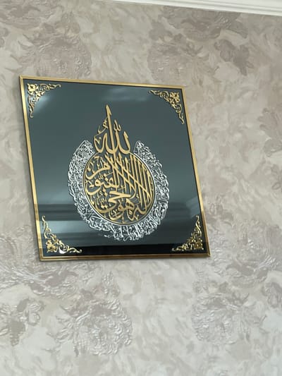 The image shows a framed piece of Islamic calligraphy hanging on a textured wallpapered wall. The calligraphy is circular in design, with gold and silver lettering against a dark, reflective background within the frame. The frame itself is gold and has decorative corner accents. The wallpaper has a raised floral pattern in lighter and darker shades of beige. The scene appears to be an indoor setting, likely a home, given the wallpaper and artwork. There are no people or specific city landmarks visible. The location in Sumgayit, Azerbaijan, is an assumption and cannot be verified from the image alone.