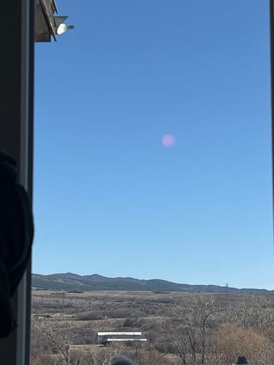 The image depicts an expansive outdoor scene viewed through a window or doorway, framing a rural landscape and sky in Golyanovtsi, Bulgaria. The upper two-thirds of the frame are dominated by a clear, bright blue sky. A distinct, diffuse, spherical pink/magenta light anomaly is centrally located in the upper-right quadrant of the sky. In the upper-left corner, a partial view of a brown, gabled architectural element, possibly a roofline or eave, is visible, from which a small, reflective, silver-colored object extends outwards. The left and right edges of the photograph show portions of a light-colored vertical frame, indicating an interior vantage point.
Below the sky, the landscape consists of undulating terrain covered predominantly with bare deciduous trees and sparse, dry vegetation, suggesting a late winter or early spring season. In the midground, a white, single-story building with a dark roof is visible amidst the vegetation. Further towards the right midground, a tall, slender telecommunication tower stands against the distant horizon. The background features a range of low, forested hills or mountains with darker patches of evergreen trees. No discernible human figures or explicit actions are present within the frame. The scene is illuminated by bright daylight.