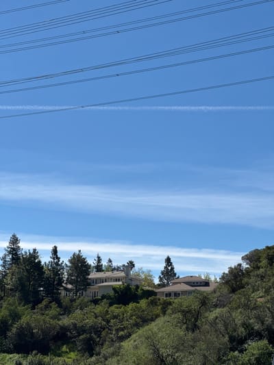 The image displays a daytime landscape in La Cañada Flintridge, United States of America. The upper two-thirds of the frame present a clear blue sky, traversed horizontally by five dark, parallel utility lines in the top section. A single, distinct white linear contrail is visible horizontally below these lines, with faint, wispy horizontal cloud formations lower in the sky. The bottom third of the image is occupied by densely vegetated hillsides, comprising various green trees and shrubs. Multiple light-colored, multi-story residential buildings are situated among the trees on a distant slope. One such residence on the left features a visible light-colored chimney structure, and a palm tree is present near another property. No human figures, animals, or specific activities are depicted.