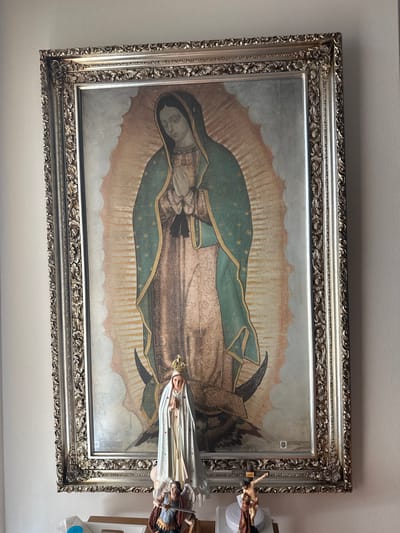 A large, vertically oriented framed print of Our Lady of Guadalupe hangs centrally on a light-colored interior wall. The ornate silver-toned frame encloses a depiction of the Virgin Mary with clasped hands, a green mantle adorned with stars, and a pink gown, standing on a crescent moon with emanating sun rays. Directly beneath the print, on a light-colored surface, are three religious figurines. Centrally positioned is a statue of the Virgin Mary, crowned, robed in white and light blue, with hands clasped. To the left, a statue of Saint Michael the Archangel is partially visible, depicted with armor, a sword, and wings, standing over a prostrate figure. To the right, a crucifix featuring Christ on the cross is partially visible. A white rectangular object rests on the surface to the far left. The scene is located indoors within the city of McAllen, United States of America.