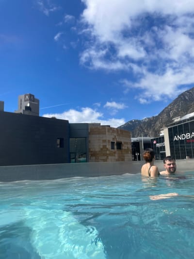 A horizontal photograph captures a scene at the Caldea Spa in Les Escaldes, Andorra. In the foreground, two individuals are partially submerged in a pool of clear, blue water, facing toward the right of the frame. The person on the left has dark hair, while the one on the right has darker features with facial hair.
To the left, the modern architecture of the building is visible, featuring geometric shapes and dark gray and tan exteriors. Above the building, a mountainous landscape extends into the distance, meeting a blue sky dotted with white clouds. The sky is bright and clear, suggesting a sunny day. A structure labeled "ANDBANC" is visible on the building behind the people in the water. The water ripples with the motion of the people, adding depth and texture to the image.