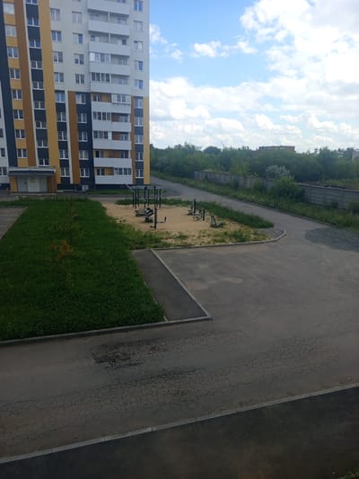 The image depicts an outdoor scene in Kopeysk, Russia. A tall, multi-story apartment building with a colorful facade, featuring sections of white, yellow, blue, and brown, dominates the left side of the frame. The building has numerous windows and balconies. Adjacent to the building is a patch of green grass, followed by a sandy area with outdoor exercise equipment, including what appears to be a pull-up bar and other metallic structures. Beyond this recreational area, a paved pathway or road, showing signs of cracking, curves away. In the background, a line of lush green trees and a grey concrete fence are visible. The sky above is a bright blue with scattered white clouds, suggesting fair weather. The perspective is from a slightly elevated viewpoint, looking down and across the scene. There is no visible text in the image. The time of day appears to be daytime, likely late morning or afternoon, given the lighting. The scene captures a residential area with amenities for outdoor activity.