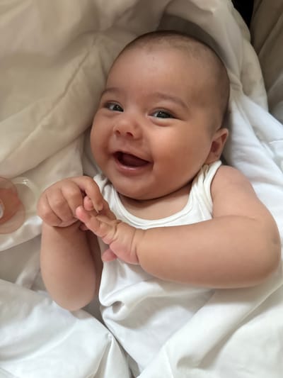 A baby is lying on a white blanket, viewed from above. The baby is wearing a white onesie and is looking towards the camera with a wide smile, mouth open and eyes bright. The baby's right arm is bent and resting on its chest, while its left arm is also bent and its hand is touching its mouth. A pacifier is visible near the bottom of the frame. The lighting is soft and natural, suggesting it is daytime, likely indoors. The background is a soft white blanket, creating a simple and uncluttered setting. The overall emotion conveyed is joy and contentment, with the baby appearing to be in a relaxed and happy state. There is no visible text in the image. The location context of Oldham, United Kingdom is noted but not visually represented in the image itself.