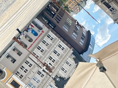 The image presents a tilted, dynamic view of an urban square or street, blending historical and modern architecture under a bright, partly cloudy sky. A large, beige outdoor umbrella or awning occupies the upper left foreground, partially framing the scene.
On the right, a multi-story building features a light gray, classically styled facade with intricate architectural details, including decorative window surrounds and cornices. It has numerous evenly spaced rectangular windows with white frames. The ground floor of this building houses a business with a blue arched entrance and displays the prominent text "VŠEOBECNÁ ZDRAVOTNÍ POJIŠŤOVNA" (General Health Insurance Company) and a visible "A" logo, along with partial "ČESKÁ" text, confirming a Czech location.
To the left, a modern, curved building rises with dark brown or gray paneled walls and large, tinted rectangular windows. Its top section features a glass curtain wall. Further in the background, glimpses of other traditional buildings with gabled roofs are visible.
An ornate, dark-colored lamppost with multiple globes stands prominently in the mid-ground, extending diagonally from the lower right. The ground is paved with light-colored stones, laid in a linear pattern, indicating a pedestrian area.
Several people are visible in the square. A woman with blonde hair, dressed in a striped top and dark pants, walks across the paved area. A small group, including a man in a blue shirt and another person in red, gathers near the entrance of the health insurance building.
The sky is a clear blue with scattered white, fluffy clouds, suggesting a sunny daytime. A red and white flag, possibly the Czech flag, is visible on a pole against the distant sky. The scene captures typical daily activity in what appears to be a lively urban center in Liberec, Czechia.