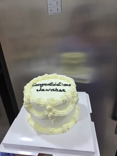 A two-tiered white cake with yellow frosting decorations is displayed on a white cake board. Black icing spells out "congratulations Jawahar" on the top tier. The cake is positioned in front of a stainless steel appliance, possibly a refrigerator, which has a control panel with icons for "ECO," "Alarm," a power symbol, and a lock symbol. The background is out of focus, but hints of a domestic or commercial setting are visible. The location is not specified in the image itself, but the question asks for it to be set in Dibba Province, Oman.