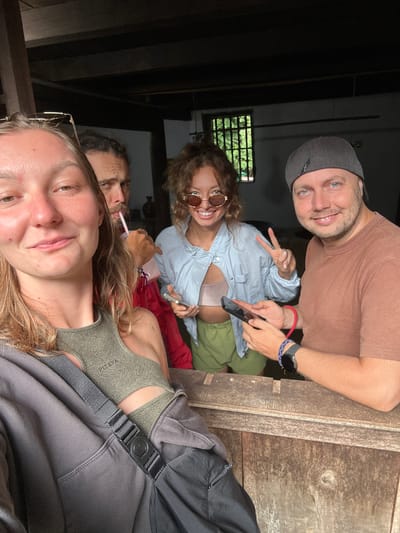 In Sà Phìn Commune, Vietnam, a group of four people are in what appears to be a rustic, dimly lit interior, possibly a wooden structure or an old building. The photo is taken from a low angle, focusing on the faces of the individuals. The person closest to the camera, likely taking the selfie, is a woman with light hair and eyeglasses. Behind her is another person drinking from a cup with a straw. Further back, a woman with curly hair and sunglasses is making a peace sign while holding a phone. A man in a cap and a brown shirt is looking at the camera with a smile. The environment suggests an informal gathering or a stop during travel. There's a wooden paneling visible in the background. The lighting is dim, emphasizing the interior setting.