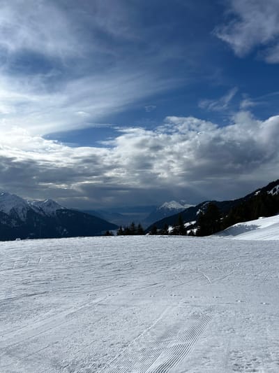 The image displays a panoramic winter landscape from a snow-covered elevation in Villard-sur-Doron, France. The foreground consists of a wide expanse of groomed snow, showing parallel tracks from a snowcat on the lower right, alongside various ski and snowboard trails. No individuals or specific objects are present on the snow.
Beyond the immediate foreground, a line of dark green conifer trees marks the edge of a midground slope, which is also covered in snow. The trees cast shadows onto the pristine white surface. In the background, a vast valley stretches out, partially obscured by atmospheric haze or low clouds. Distant, rugged mountain ranges with snow-capped peaks are visible on both the left and right sides of the valley. The sky above is a mix of bright blue patches and various cloud formations, ranging from high, wispy cirrus clouds to lower, denser cumulus and stratocumulus clouds, casting dynamic light and shadows across the scene. The overall lighting suggests partial sunlight breaking through the cloud cover.