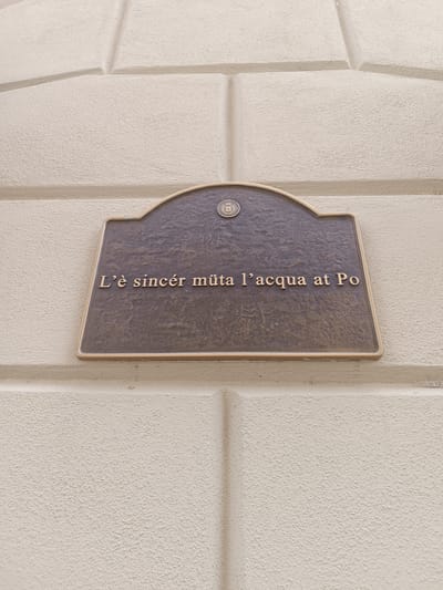 A bronze plaque with an arched top is mounted on a light-colored, textured wall. The plaque displays embossed text in a serif font. The text reads: "L'è sincer mûta l'acqua at Po". At the top center of the plaque, a circular emblem with a crest is visible. The wall itself has a pattern of recessed horizontal and diagonal lines, resembling stone blocks. The lighting suggests an outdoor setting, likely during the daytime. This plaque is located in the city of Casalmaggiore, Italy.
