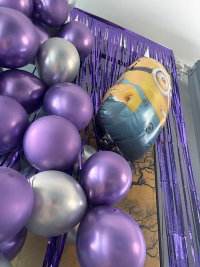 A close-up, low-angle shot shows a collection of metallic purple and silver balloons, along with a large foil balloon shaped like a Minion character, arranged as a party decoration. The balloons are clustered together, obscuring much of the background. Behind the balloons, a shimmering purple fringe curtain hangs vertically, suggesting a festive backdrop. To the right of the fringe, a portion of a gray curtain is visible. The balloons are the primary focus, with the Minion balloon positioned prominently among them, facing to the right. The overall scene appears to be indoors, possibly set up for a celebration. The location of Natukhayevskaya, Russia, is not depicted in the image.