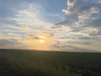 The image depicts a rural landscape at sunset, likely in the vicinity of Meden Kladenets, Bulgaria. The foreground features a field of green vegetation, possibly crops or grasses. The horizon line is slightly above the center of the frame, with faint hills or mountains visible in the distance. The sky is filled with a mix of blue and gray clouds, illuminated by the setting sun. The sun itself is partially obscured by clouds, creating a diffused golden light. No people or animals are visible. The overall scene suggests a tranquil, agricultural setting.