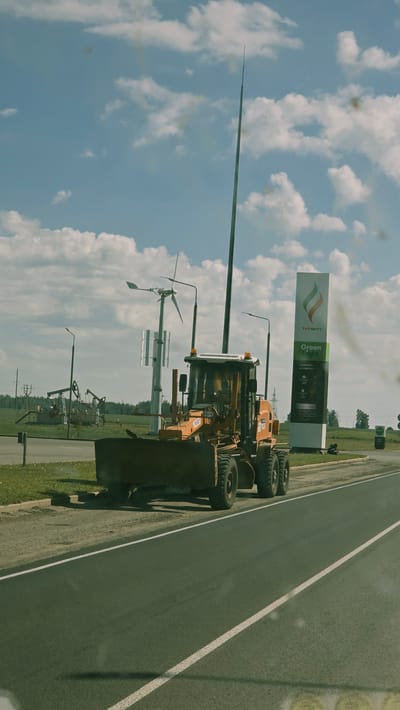 An orange grader is parked on the shoulder of a paved road. The grader has a large blade at the front and large tires. Behind the grader, to the left, are several oil derricks and a small wind turbine. To the right of the grader is a tall, slender pole topped with a pointed object. Further to the right is a large vertical sign for "TATNEFT Green Light" which appears to be a gas station. The sky above is blue with scattered white clouds. The scene appears to be in a rural or industrial area.