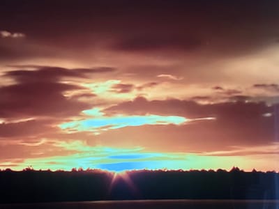 This media file captures a sunset over a silhouetted treeline, likely on the outskirts of Valladolid, Spain. The sky is a dramatic display of color, with hues of deep purple, fiery orange, and bright yellow. Wispy clouds are scattered across the sky, catching the last rays of the sun and creating a layered effect. A band of lighter, almost turquoise light is visible just above the treeline, indicating the sun is setting or has just set below the horizon. The treeline itself is dark and irregular, hinting at a forest or dense vegetation. The overall mood is serene and awe-inspiring, characteristic of a beautiful sunset. There is no visible text in the image.