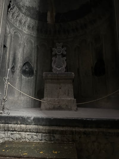This image captures the dimly lit interior of a rock-cut chamber within the Geghard Monastery complex, situated near the village of Goght, Kotayk Province, Armenia. A prominent stone monument stands centrally, featuring a large, intricately carved cross surmounting a multi-tiered rectangular pedestal. The cross itself displays elaborate scrollwork and geometric patterns. The surrounding walls, carved from dark, textured stone, are adorned with architectural reliefs, including vertical fluted elements, arched recessed panels, and two deep, dark, arch-shaped niches—one visible on the left wall and another on the right wall, equidistant from the central monument. Above these, a decorative frieze of repeating pointed arch motifs follows the curve of the chamber's upper section. In the foreground, a low stone barrier or step separates the viewer's space from the monument. Below this step, a shallow, rectangular pool of dark water contains scattered yellow-orange flecks, possibly offerings. A light-colored rope, attached to a decorative metal stanchion on the left, stretches horizontally across the lower part of the scene, acting as a visitor barrier. No individuals are present in the frame. The lighting is low and uniform, emphasizing the ancient, monochrome stone.