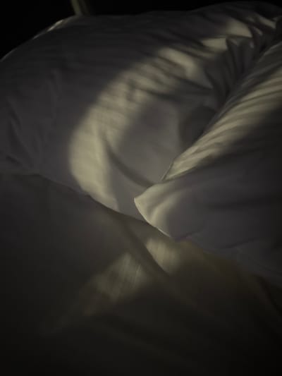 The image depicts a close-up view of a white duvet or comforter with subtle striped patterns. A diagonal shaft of light illuminates the textured surface, creating areas of highlight and shadow. The lighting suggests the presence of natural light, possibly from a window, casting distinct linear shadows across the fabric. The overall impression is one of a peaceful, undisturbed bed. The location information provided, "Doma 1096 Km, Russia," appears to be extraneous and does not correspond to any discernible elements within the image itself.