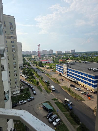 This is a high-angle, medium shot of an urban scene on a partly cloudy day. The image captures a view from a balcony overlooking a street lined with residential buildings, a large commercial building, and distant city structures.
In the foreground on the left is the side of a light-colored residential building, with several windows visible. A white metal railing runs along the bottom of the frame.
Dominating the middle ground is a road curving through the scene, with several cars parked along its sides and others driving. To the left of the road, a light green multi-story residential building is prominent, with balconies and windows. Adjacent to this building is a parking lot filled with cars.
Across the street, a large, multi-story commercial building with a distinctive blue and white striped facade stands out. Several cars are parked in front of this building, and a few individuals are walking on the sidewalk.
Further in the background, the landscape transitions to more residential buildings and industrial structures. Tall red and white striped smokestacks are visible in the distance, as well as numerous apartment blocks and green trees. The sky is a mix of blue and white clouds.
The scene depicts a typical urban environment with elements of residential living, commerce, and infrastructure. The overall impression is one of a moderately busy city on a pleasant day.