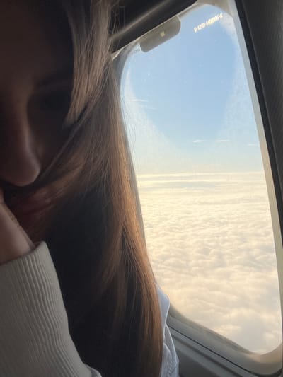 This is a photograph taken from inside an aircraft, looking out the window. The foreground is dominated by the back of a person's head and long, brown hair, which falls across the frame. A white sleeve of clothing is also visible on the right side of the image.

Outside the window, a vast expanse of sky is visible. The sky is a clear blue on the left, transitioning to a sky filled with soft, fluffy white clouds on the right, suggesting either dawn or dusk. Faint contrails from other aircraft can be seen in the blue sky. The setting is a flight, likely in the early morning or late afternoon, judging by the light. There is no discernible emotion conveyed, as the focus is on the view and the person's hair.

On the aircraft window, there is some faint text. It appears to be a series of numbers and letters, possibly a part number or identification. The text reads: "B-1210-1406506-K". The location context is Stolbishche, Russia.