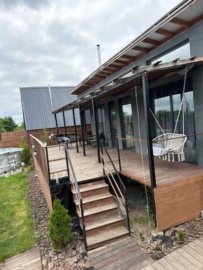 The image depicts the exterior of a modern house with a wooden deck and a covered porch in the city of Semiluki, Russia. The house has a gray facade and large windows. On the deck, there is a white hanging swing chair with cushions, several chairs and tables, and string lights. Stairs lead down from the deck to a wooden walkway. To the left of the deck, a small above-ground pool is visible, surrounded by grass and bark mulch. In the background, another building with a gray, corrugated metal roof is present. The sky is overcast with gray clouds. The overall scene suggests a residential property, possibly a vacation home or a modern dwelling.