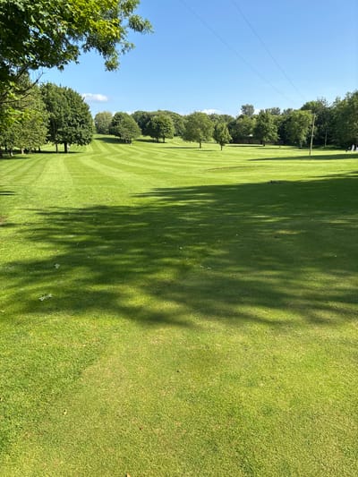 The image shows a golf course in Lancaster, United Kingdom, located at 54.01 latitude and -2.829 longitude. The green grass is neatly mowed, and there are several trees lining the edge of the fairway. Long shadows cast by the trees stretch across the course. The sky is blue with white clouds and several power lines run across the scene. The grass in the foreground is a light green, while the grass in the background is a darker green. There are no people or other objects in the image, except for the trees and the power lines. The photo was taken from the perspective of a golfer on the fairway, looking down towards the green.