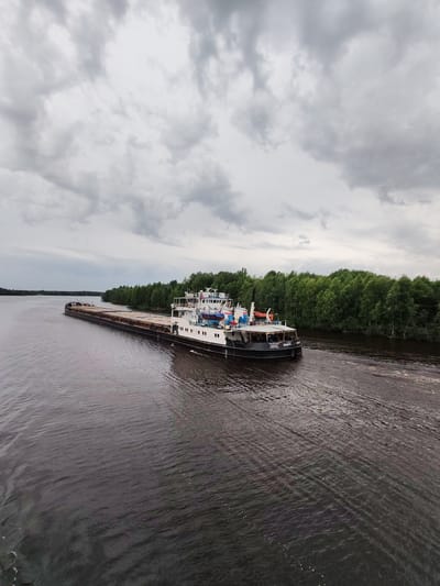 A large barge, laden with cargo, travels down a wide, dark river. The barge is long and rectangular, with a dark hull and a lighter deck carrying what appears to be timber or other building materials. A white superstructure sits at the stern of the barge, housing the vessel's bridge and living quarters. A smaller tugboat is pushing the barge, also visible at the stern.
The riverbanks are lined with a dense forest of green trees, extending as far as the eye can see. The water is calm, with gentle ripples reflecting the overcast sky. The sky itself is filled with heavy, grey clouds, suggesting a subdued or potentially rainy day. The overall scene is one of quiet industry and natural beauty, typical of a waterway in a forested region.
Visible text on the stern of the tugboat reads "ВОЛГО - DON". The scene is likely taking place in the daytime, given the natural light. The location is identified as Novokemskii, Russia, consistent with the type of waterway and the vessel markings. No people are clearly visible, but their presence is implied by the operation of the tugboat and barge.