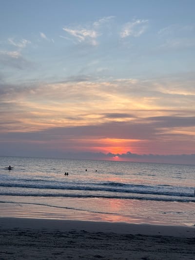 A sunset at the beach features an array of clouds painted in hues of orange, yellow, and blue. Several people are visible in the ocean, some swimming, and some standing. The water reflects the warm colors of the setting sun. The sandy beach stretches along the right side of the image.