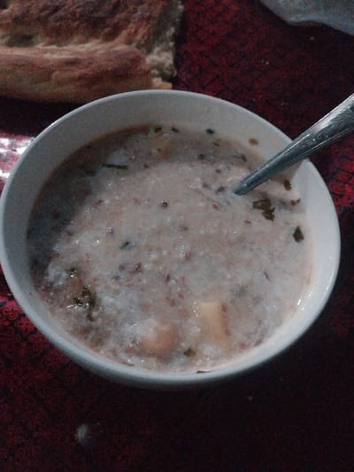 A close-up, overhead shot shows a white ceramic bowl of porridge on a dark red, patterned tablecloth. A silver spoon rests in the bowl, partially submerged. The porridge is thick and creamy, with visible grains and small green flecks. In the upper left corner, a piece of flatbread is visible, slightly out of focus. The scene is set indoors, possibly in a home in Vahdat, Tajikistan, as indicated by the style of the bread and the overall presentation. No people are visible in the image.