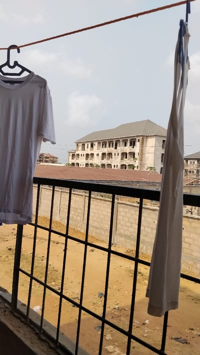 The image displays an outdoor urban environment in Isieke, Nigeria, observed from an elevated vantage point. In the immediate foreground, an orange clothesline spans horizontally across the upper frame. A white short-sleeved t-shirt hangs from a black plastic hanger on the left side of the clothesline, while a white sleeveless garment hangs vertically from the clothesline on the right. Below the hanging laundry, a black metal grid-like railing or grille is present.
Beyond the railing, the ground is composed of reddish-brown earth with scattered debris, including white plastic fragments. A light grey concrete block wall, topped with barbed or razor wire, extends horizontally across the midground. Behind this wall, a structure with a reddish-brown corrugated metal roof is visible.
In the background, a large, unfinished multi-story concrete building stands prominent. This structure features multiple levels with numerous open rectangular window and door apertures and an uncompleted exterior, topped with a grey hip roof. A second, smaller unfinished building is partially visible to the right. The sky is light blue with scattered white clouds. No individuals are depicted.