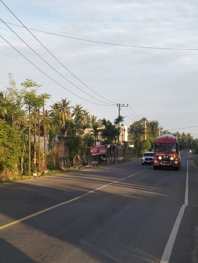 The image shows a road in Teungkluet, Indonesia, with a red truck and a white car driving on it. On the left side of the road, there are trees and a small shack with advertisements on it. There are also power lines running across the sky. The sky is light blue with some clouds. The road has a yellow line in the center and a white line on the right edge.