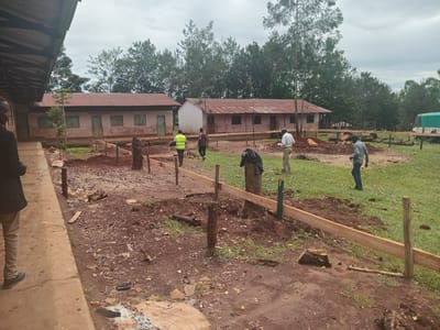 The image depicts an outdoor scene in Idakho South ward, Kenya, with construction or renovation work underway. Two long, low-rise buildings with rusty, corrugated metal roofs and beige or light-colored walls stand in the background. A group of at least five individuals is scattered across the foreground and middle ground. One man is visible on a sidewalk or path on the left edge of the frame. Another person, wearing a reflective vest, walks near the buildings in the middle distance. A man is visible holding papers. Another is standing near the grass and a truck. There are wooden stakes and boards arranged as if to create forms for concrete work. The ground is dirt and sparse grass. The sky is overcast. Trees are visible behind the buildings. A truck is visible to the right of the buildings.
