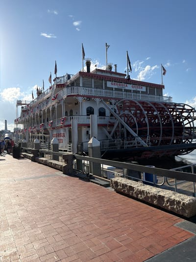 The image shows the Georgia Queen sternwheeler docked at River Street in Savannah, Georgia. The large paddlewheel boat is white with red trim and decorated with American flags and red, white, and blue bunting. The boat has multiple decks with railings and windows. The stern of the boat features a large, red and white paddlewheel. The foreground shows a brick walkway leading to the dock. The dock has a metal railing and stone benches. Several people are visible walking on the walkway. The sky is blue with scattered clouds. The overall scene depicts a sunny day at a popular tourist location.