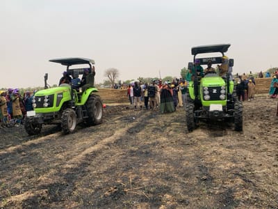 The image shows two green tractors, both with the word "WOFAN" written on the side, positioned in a field of burnt grass near Ringimawa, Nigeria, at the coordinates 11.666 latitude and 8.416 longitude. The tractor on the left is facing the right, and the tractor on the right is facing the left. The tractor on the left has a canopy and three individuals are seated inside. The tractor on the right has a canopy with three individuals inside. There is a group of approximately 50 people standing in the background, all wearing traditional clothing. A large speaker is visible in the background between the tractors. The scene is located in a field with burnt patches of grass and visible tire tracks. The sky is a light blue color with scattered white clouds.