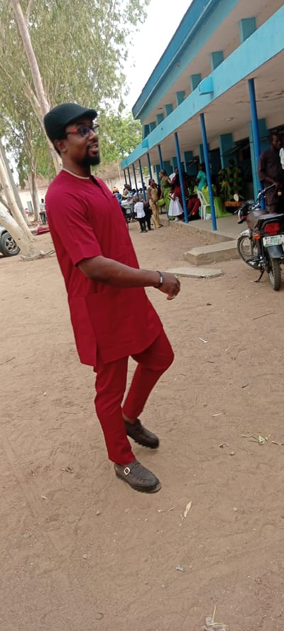 A man is standing outdoors in Goron Dutse, Nigeria. He is wearing a dark cap, glasses, and a red traditional outfit consisting of a short-sleeved tunic and trousers. He is wearing brown buckled shoes. The ground is sandy. Behind him is a single-story building with a blue roof and blue columns supporting a covered walkway. People are seated and standing under the walkway. A motorcycle is parked on the right side of the image. Trees are visible in the background. A car can be seen on the left side of the image.