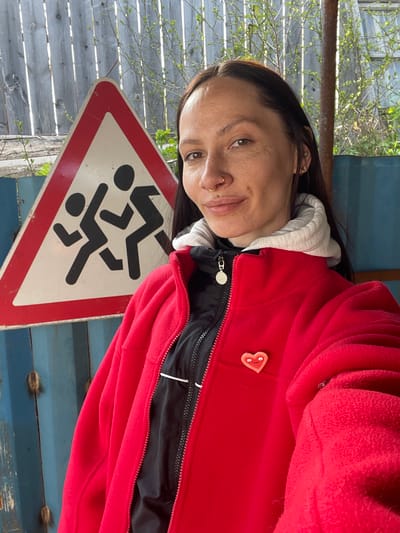A woman with brown hair and fair skin stands next to a triangular warning sign with a red border, depicting two stick figures running, presumably a children's crossing sign. She is wearing a red fleece jacket over a gray hoodie and a black zip-up inner layer. A heart-shaped button with a smiling face is attached to the jacket. The scene is set against a background of weathered wooden fences and a blue metal container. The location is likely near Doma 45 Kilometr, Russia, due to the context provided. The woman is looking at the camera.