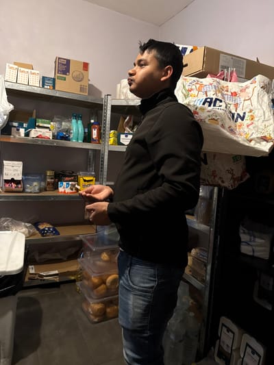 One male individual is depicted in a storage or pantry area located in Holtzheim, France. The subject, appearing to be of South Asian or Southeast Asian descent, stands in a right-side profile, looking upward and slightly to his right. He is wearing a black long-sleeved turtleneck jacket and distressed blue jeans. Both hands are positioned in front of his torso, holding a small, dark, slender object, with his left hand cupping the object and his right hand partially visible.

The environment consists of two metal shelving units lining light-colored walls (pale purple/grey) with a dark tiled floor. The shelving units are laden with various items:
*   **Left shelving unit:** Contains multiple boxes, including a "TORK" brand paper towel dispenser box. Below are plastic bottles, likely cleaning supplies, alongside various packaged goods, small containers, and snack items such as a "Nescafe" jar, a "Mars" bar, and a "Kinder" product. Two calendar-like items are visible on the top shelf.
*   **Right shelving unit:** Features more stacked boxes on the top shelf, partially covered by a large, crumpled white bag bearing the "ACTION" logo and colorful illustrations. Lower shelves contain clear plastic containers stacked with numerous bread rolls or buns. Several large plastic bottles, possibly containing water, are on the floor next to this unit, along with white bags, possibly containing dry goods.

A white plastic bin with a dark liner is partially visible on the far left. The lighting in the room appears to be overhead, casting some shadows. No other individuals are present in the frame.