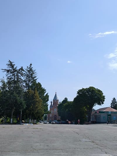 The image captures a wide-angle view of a public square or open area in Illintsi, Ukraine, under a clear, bright blue sky with a few wispy white clouds. The foreground consists of an expansive, light grey asphalt surface exhibiting cracks. In the midground, the prominent subject is a large, multi-domed church constructed of red brick, featuring a central spire topped with a cross. This church is partially framed by various trees, including tall, slender coniferous trees on the left and a mix of deciduous and coniferous trees in the center. A flagpole bearing a yellow and blue flag is visible to the left of the church. Several vehicles are parked in front of the church. To the right of the central view, a large, full deciduous tree partially obscures a single-story commercial building with a green roof. This building is identified as a pharmacy by Ukrainian text "АПТЕКА ПОДОРОЖНИК" on its facade. Three to five indistinct individuals are present in the midground; one person wearing a red top stands near the large deciduous tree on the right, and another individual appears to be bending near the parked cars. A scooter or motorcycle is visible adjacent to the pharmacy building. The scene indicates daylight conditions.