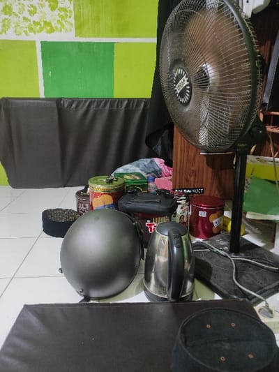 The image shows a cluttered scene, likely inside a home or a small business. The location is Peterongan, Indonesia, based on the provided coordinates. The main subject of the image is a black electric fan, mounted on a black metal stand and positioned against a wall. The fan is facing the right side of the image. To the left of the fan is a black, padded seat.  The scene is cluttered with objects, including two black hats, one black helmet, a silver kettle, two red metal cans with labels, a black zippered bag, and various other items.  The wall behind the seat is painted in a green and white pattern. The floor is tiled in white and gray. The image suggests a general lack of organization in the space.  The objects and the overall clutter suggest this scene may be from a shop or home in a developing country.