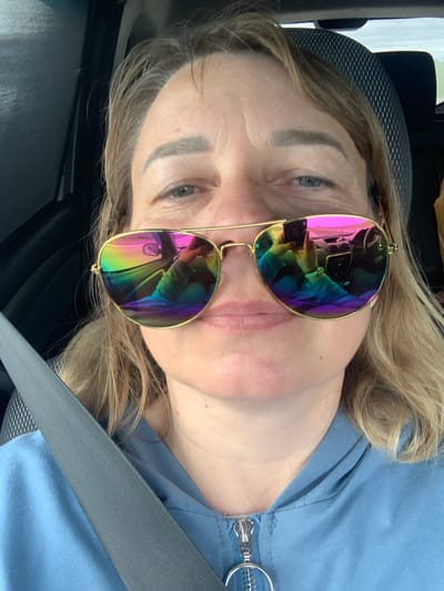 A close-up selfie shows a woman wearing sunglasses with rainbow-colored lenses. She is in the driver's seat of a car, with the car's dashboard and steering wheel reflected in her sunglasses. The woman is wearing a blue hoodie with the zipper partially down. A seatbelt is visible across her chest. The background is out of focus, but appears to be a road with some greenery. The scene is consistent with a person taking a selfie while driving in the Азерскі сельскі Савет region of Belarus.