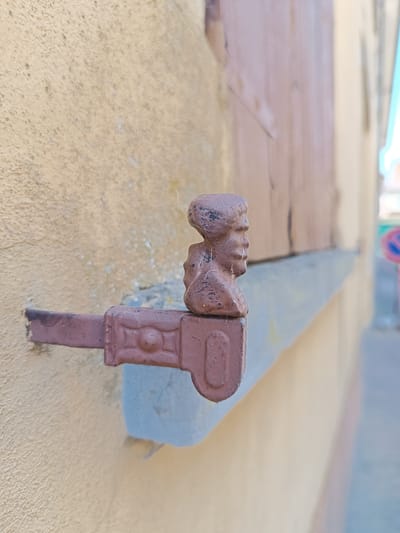 The image shows a close-up of a weathered metal window latch attached to a beige wall in Mezzana Sant'Antonio, Italy. The latch features a decorative bust profile on its upper portion. The wall's surface is textured and appears aged. The latch itself is reddish-brown and displays signs of rust and wear. In the background, there is a glimpse of a wooden shutter, a street scene with blurred details including a traffic sign, suggesting an urban environment.