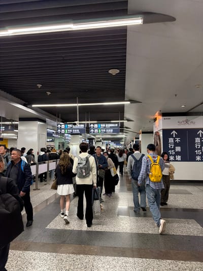 The image depicts a moderately busy underground transit station in Yuhuatai District, China. The environment is characterized by modern, functional architecture. The ceiling features dark gray slatted panels with embedded fluorescent strip lighting, interspersed with smoke detectors and fire suppression heads. Portions of the ceiling and upper walls are light gray. The flooring is a speckled light gray with dark gray rectangular pathway indicators.
Approximately two to three dozen individuals, primarily adults, are visible, engaged in transit activities. Most are walking in the direction away from the camera, though some are seen in profile or facing the camera in the mid-ground. Attire is predominantly casual, including sweaters, jackets, shirts, and jeans. Many individuals are carrying personal items such as backpacks and rolling luggage. A person in the foreground wears a white sweater and carries a light grey backpack and a patterned rolling suitcase. Another individual further back wears a plaid shirt and a bright yellow backpack.
Overhead, multiple white-on-blue directional signs with white arrows indicate routes for "1号线 Line 1" (Line 1) and "3号线 Line 3" (Line 3), specifying destinations such as "往 中国药科大学" (towards China Pharmaceutical University) and "往 林场" (towards Lincun). Along the right wall, a large informational or advertising panel displays an artistic graphic and Chinese text, including "直行 15 米" (go straight 15 meters). People are observed moving purposefully through the station, suggesting travel to or from platforms or exits. Interactions among individuals appear limited to walking in proximity.