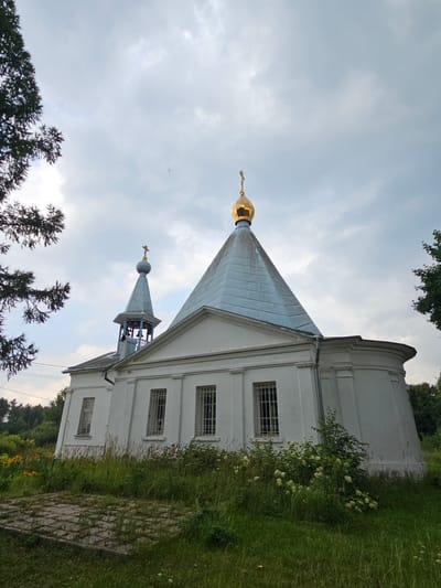 A white Orthodox church with a golden dome and a blue, conical roof is depicted from a low-angle perspective. To the left of the main building, a smaller belfry with a blue, pointed roof and bells is visible. The church has several rectangular windows with metal grates. The surrounding area is overgrown with green grass and vegetation, with some yellow flowers and bushes with white blooms. Trees frame the scene on the left and right. The sky is cloudy. The church is located in the village of Ulitkino, Russia.
