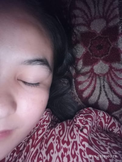 A close-up, low-angle, indoor, dimly lit, overhead shot captures a person's left eye and forehead. The individual is resting on a textured blanket with a floral pattern of red and beige. Another patterned fabric, predominantly red with white swirls, is visible beneath the person's chin and extending across the bottom right of the frame. A timestamp "30.06.2026 00:13" is overlaid on the bottom right corner. The setting is a domestic environment in Batken City, Kyrgyzstan.