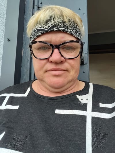 A woman with blonde hair and a bandana is pictured in an outdoor setting. She is wearing large, patterned eyeglasses and a dark t-shirt with white geometric patterns and a cat design. The background consists of a grey, textured wall and a dark-colored door or frame, possibly part of a building's exterior. The overall scene suggests a casual, everyday moment captured in front of a residential or utility structure. The image is a medium shot, focusing on the woman's face and upper body.