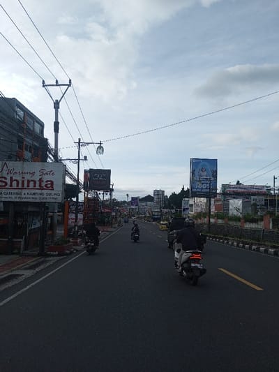 The image shows a street with several motorcycles riding on the asphalt. On the left side of the street, there is a building with a large sign that reads "Warung Sate Shinta" advertising catering and kambing guling. Further down the road, there are several other billboards and signs. The sky is overcast, and there are power lines stretching across the top of the image. A yellow line is painted on the road dividing the lanes.