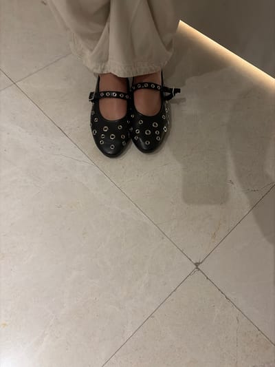 The image depicts a pair of black, round-toe Mary Jane shoes with eyelet details and ankle straps, worn by a person standing on a tiled floor. The person is wearing beige or off-white trousers. The location appears to be indoors, possibly within a building or commercial space in the city of Key Biscayne, United States of America, based on the prevalence of tiled flooring and contemporary design elements. There is no discernible interaction or action beyond the stationary position of the individual. The environment is brightly lit with some directional lighting.